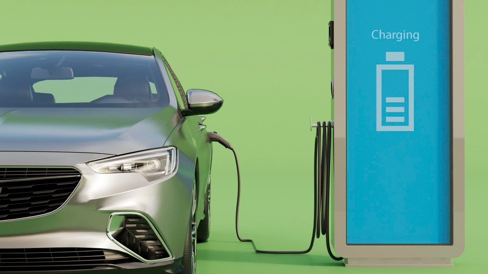 A silver car plugged into an electric charging station, with a clear view of the charging cable and station signage.