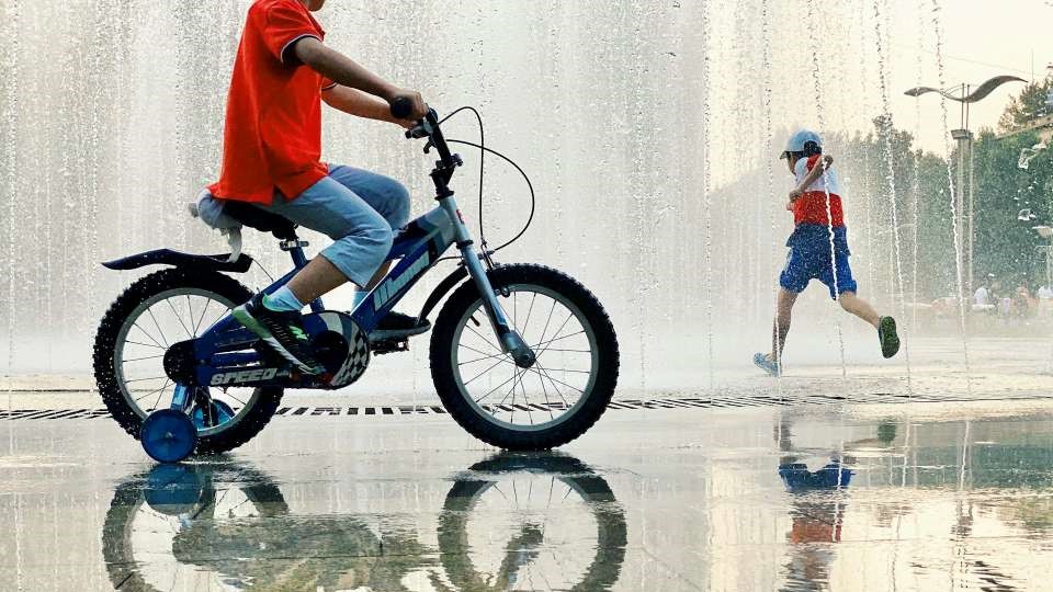 A boy on a bicycle rides in front of a fountain, with water flowing and sunlight creating a sparkling effect around him.